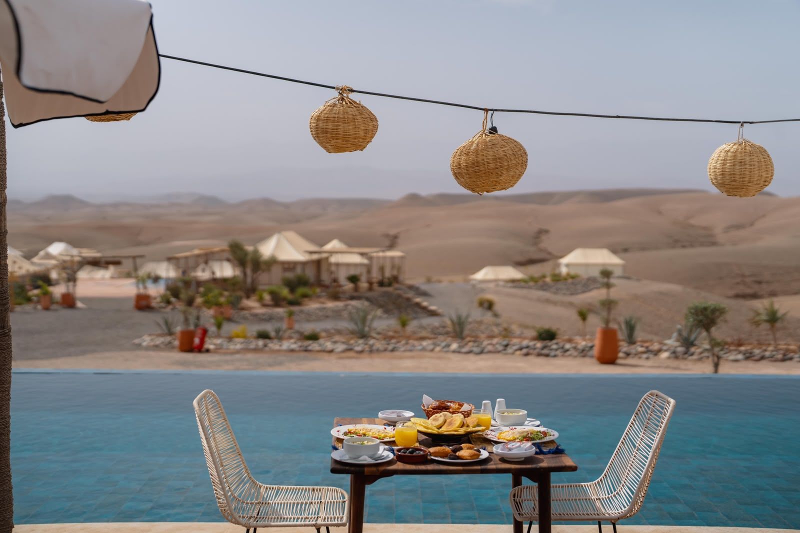 Moroccan breakfast laid out on a table beside the infinity pool, wicker lanterns and the tents of the camp beyond