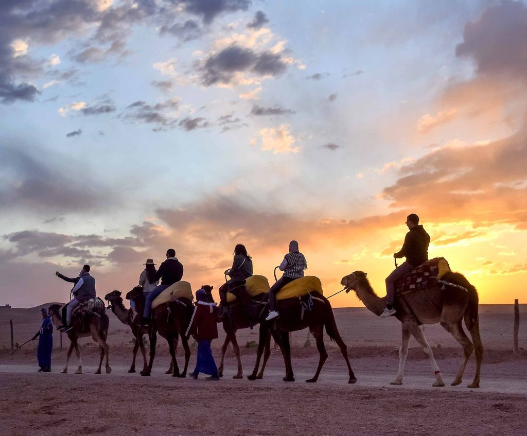Line of camels and riders crossing the Agafay Desert against an orange sunset sky