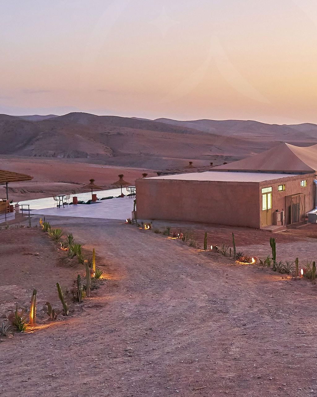 Camp buildings and infinity pool in the pink light of dusk, the Agafay hills beyond