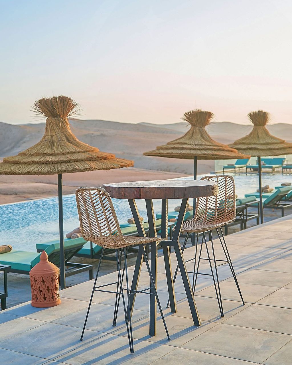 Stehtisch und geflochtene Hocker am Pool bei Sonnenuntergang, Strohschirme und Wüstenhügel dahinter