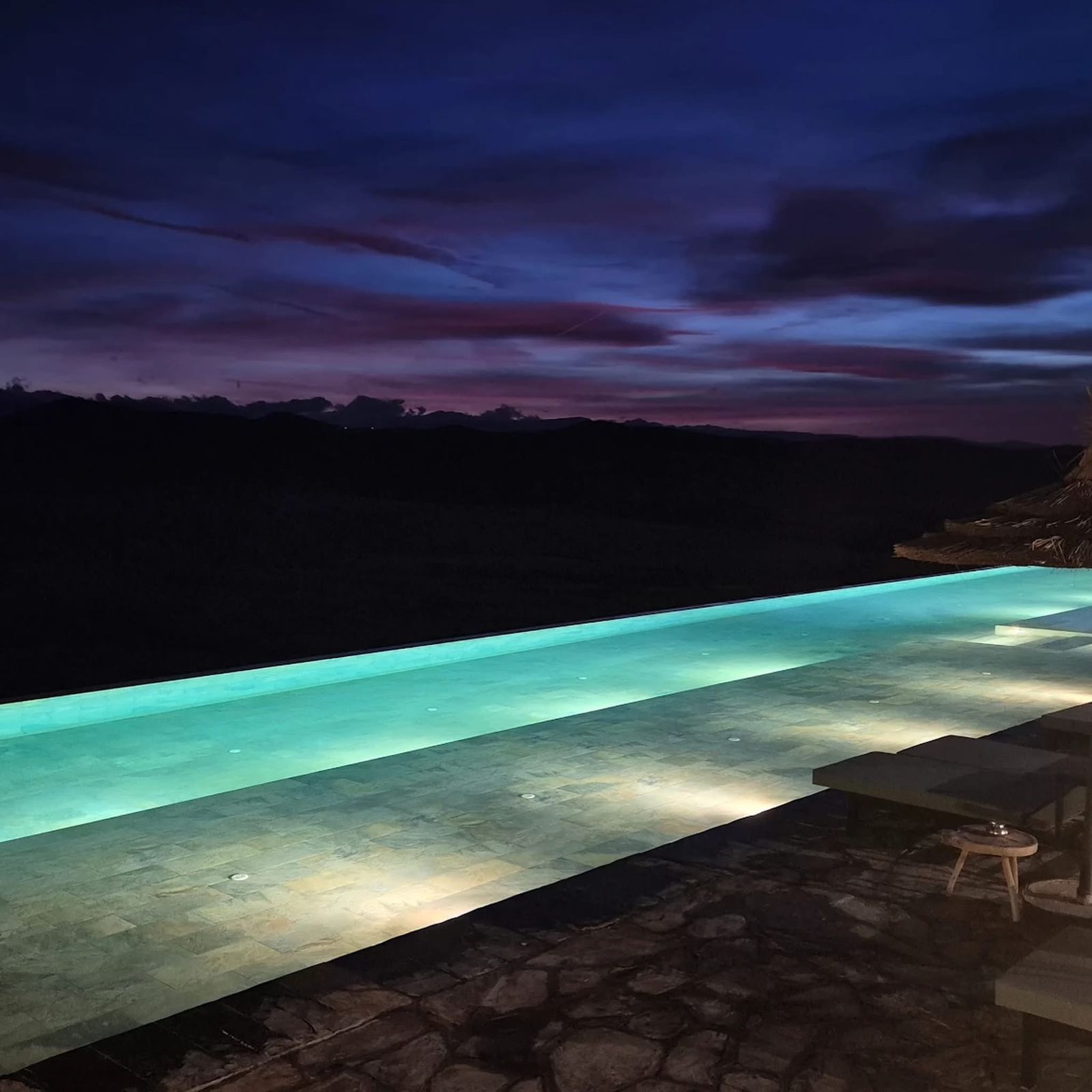 Long infinity pool lit turquoise after dark, the Agafay hills a dark line under a violet sky
