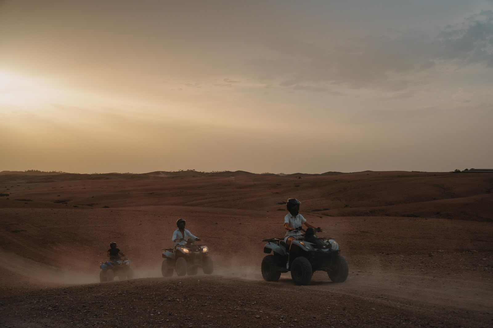 Three quad bikes crossing the Agafay plateau at sunset, raising a trail of dust