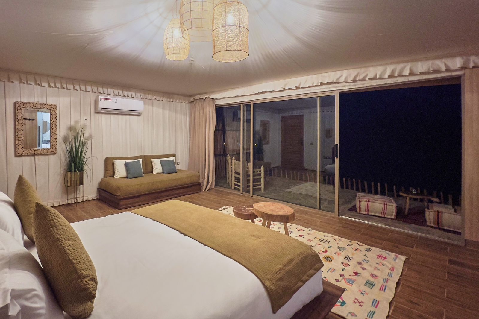 Family tent lit in the evening, with a double bed, a sofa and a terrace beyond the glass
