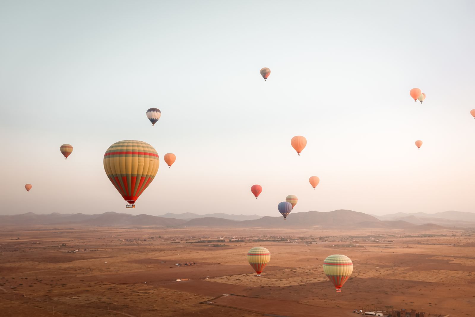 Around fifteen balloons in flight over the dry plain, with hills on the horizon
